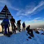 Des randonneurs au sommet enneigé du mont Toubkal, à côté du repère du sommet, avec une vue panoramique sur les montagnes de l’Atlas.