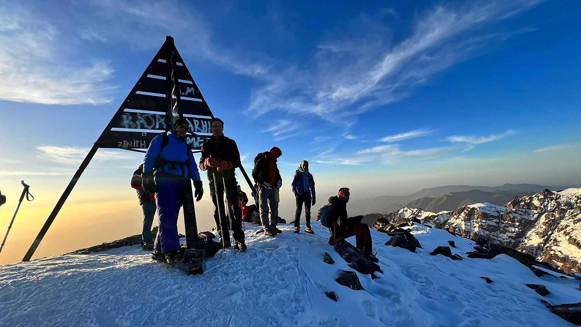 Des randonneurs au sommet enneigé du mont Toubkal, à côté du repère du sommet, avec une vue panoramique sur les montagnes de l’Atlas.