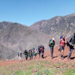 Groupe de randonneurs traversant une crête de terre rouge avec vues panoramiques sur la chaîne du Haut Atlas et des vallées profondes derrière — col d'altitude sur le trek de 3 jours Imlil à Setti Fadma