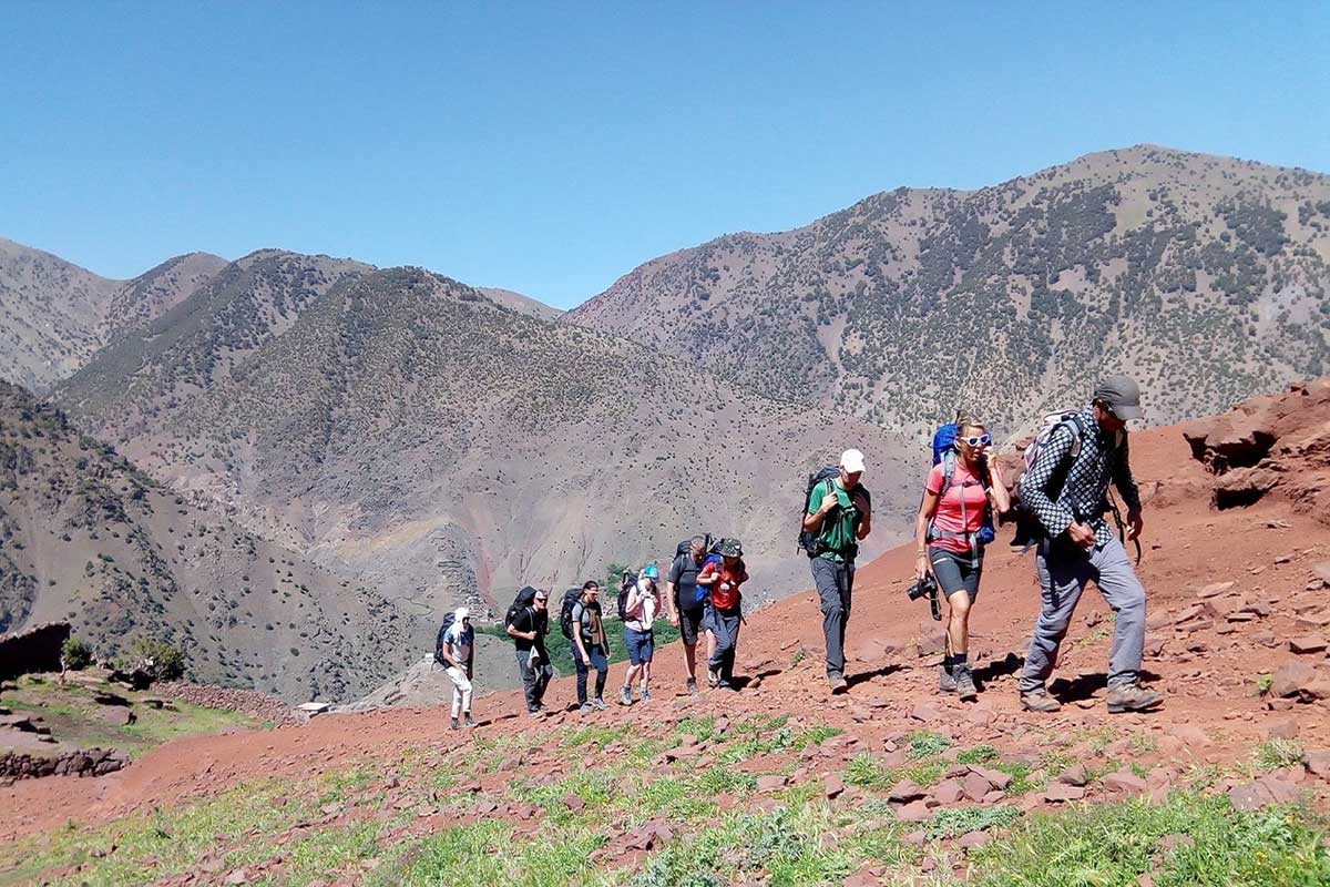 Group of trekkers crossing a red earth ridge with sweeping views of the High Atlas mountain range and deep valleys behind — high pass on the 3-day Imlil to Setti Fadma trek